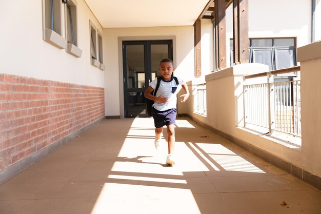 Foto longitud completa de un estudiante de primaria afroamericano con mochila corriendo en el corredor de la escuela