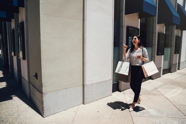 longitud completa de una joven asiática caminando con bolsas de compras fuera de la joyería en el centro comercial de stanford en venta de verano. niña alegre emocionada señalar con el dedo encontrar tienda por teléfono inteligente mapa en línea.