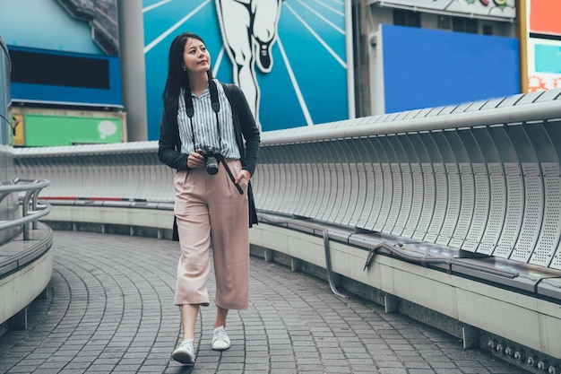 longitud completa de una joven japonesa que visita la ciudad de osaka, japón, el fin de semana. mujer caminando sobre el puente dotonbori llevando una cámara profesional mirando a un lado sonriendo. hermoso turismo turístico al aire libre.