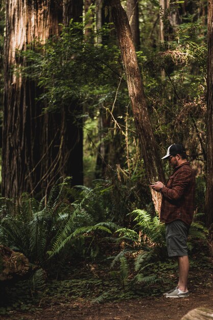 Foto longitud completa de un joven de pie junto a un árbol en el bosque operando un control remoto de avión no tripulado mirando hacia arriba