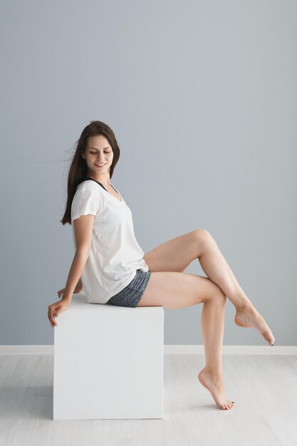Foto la longitud completa de la mujer joven sentada en el bloque blanco contra la pared gris en casa