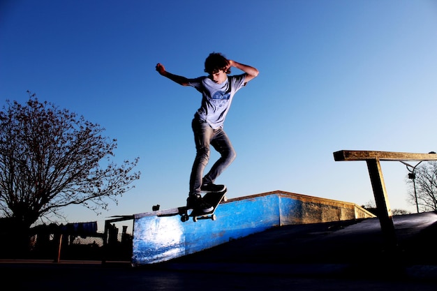 La longitud completa del niño patinando contra el cielo azul claro durante la puesta de sol
