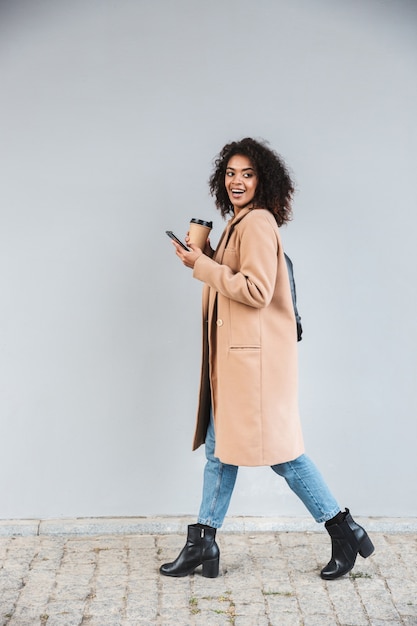 Longitud total de una alegre joven africana vistiendo abrigo caminando al aire libre, sosteniendo una taza de café para llevar, usando el teléfono móvil