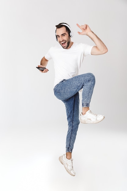 Foto longitud total de un apuesto hombre alegre con camiseta en blanco que se encuentran aisladas sobre una pared blanca, escuchando música con auriculares, usando el teléfono móvil