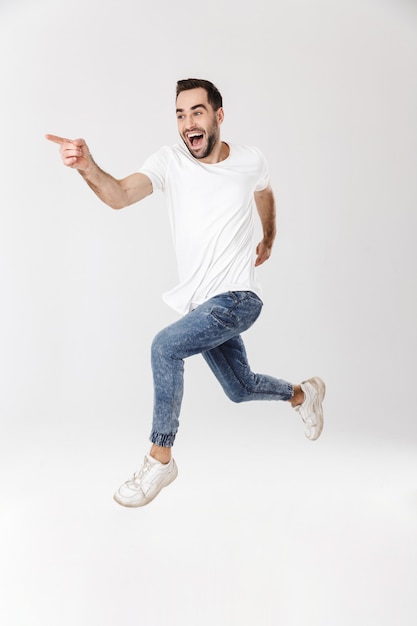 Longitud total de un apuesto hombre alegre con camiseta en blanco que se encuentran aisladas sobre una pared blanca, señalando con el dedo, saltando