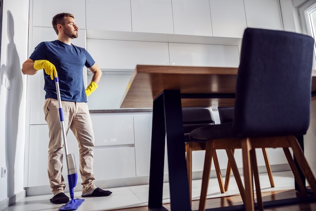 Longitud total de apuesto hombre cansado de pie en la cocina y apoyado en un trapeador. Hay mucho que limpiar.