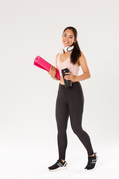 Longitud total de la atleta femenina asiática delgada y hermosa sonriente, la muchacha lleva la estera de goma del ejercicio físico y la botella de agua, yendo a clases de yoga o entrenamiento en el gimnasio con equipo propio, fondo blanco.