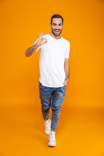 Longitud total de chico guapo en camiseta y jeans mostrando signo de ok mientras está de pie, aislado en amarillo