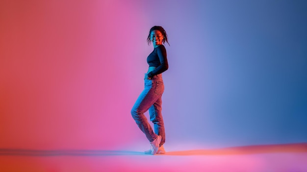 Longitud total de elegante mujer afroamericana posando con luz de neón en el estudio y sonriendo a la cámara