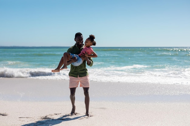 Longitud total de feliz hombre afroamericano con hija en la playa contra el cielo en un día soleado