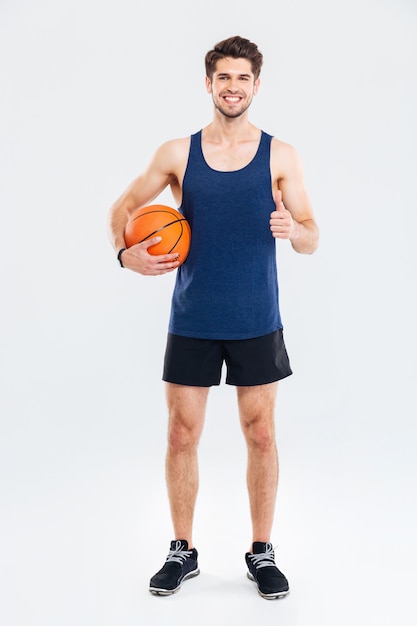 Longitud total de feliz joven deportista sosteniendo una pelota de baloncesto y mostrando los pulgares hacia arriba sobre fondo blanco.