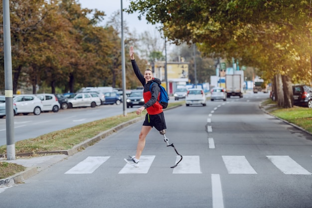 Foto longitud total de guapo deportista discapacitado caucásico en ropa deportiva, con pierna artificial y mochila cruzando la calle y saludando a un amigo.