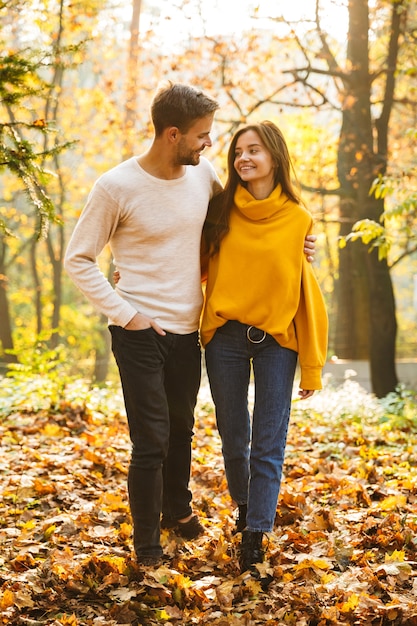 Longitud total de una hermosa joven pareja de enamorados caminando en el parque de otoño, tomados de la mano