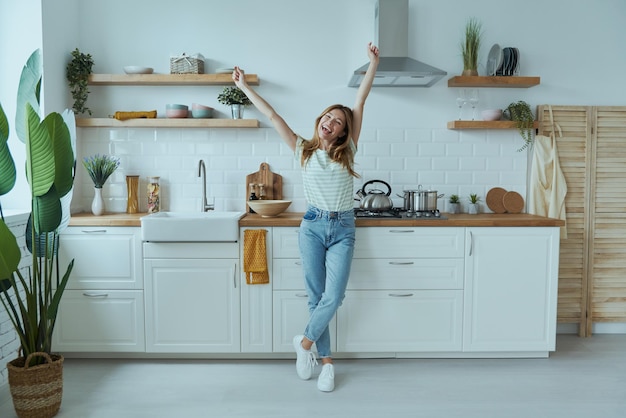 Longitud total de una hermosa joven que se ve feliz mientras está de pie en la cocina doméstica