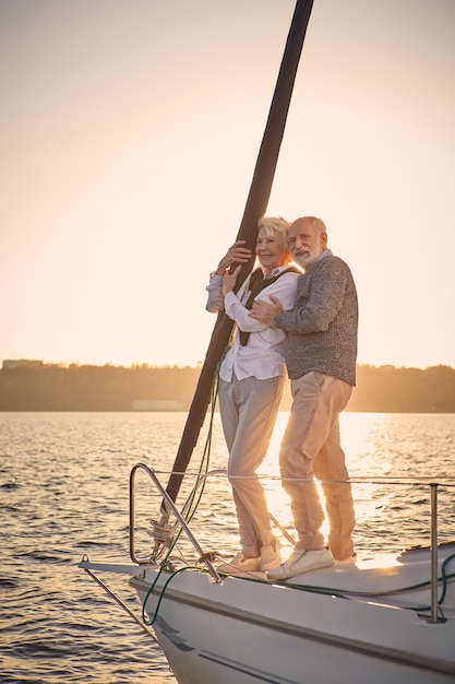 Longitud total de la hermosa pareja senior feliz en el amor de pie al lado del barco de vela flotando en