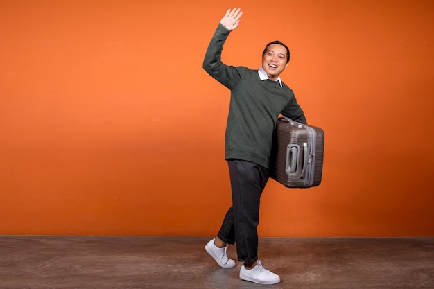 Longitud total del hombre asiático saludando con la mano diciendo hola o adiós mientras sostiene el equipaje