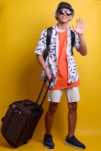 Longitud total de un hombre asiático vestido con ropa de playa agitando la mano diciendo hola o adiós mientras sostiene el equipaje