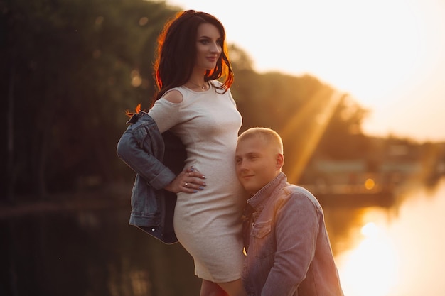 Foto longitud total de hombre encantado con chaqueta de mezclilla escuchando y abrazando a su futuro bebé dentro del vientre de su esposa están en un hermoso parque junto al río o al lago al atardecer