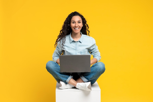 Longitud total de una joven alegre sentada con las piernas cruzadas con una laptop sonriendo a la cámara trabajando o