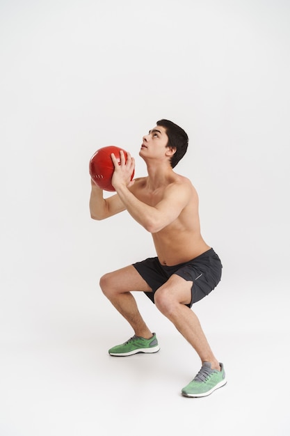 Longitud total de un joven deportista sin camisa en forma segura haciendo ejercicios con pelota pesada sobre blanco