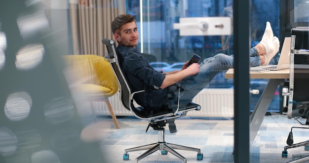 Longitud total de un joven empresario casual relajado sentado con las piernas en el escritorio en la oficina