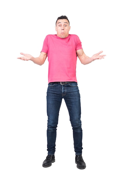 Foto longitud total de joven incierto en camiseta y jeans encogiéndose de hombros y haciendo expresión de cara confusa contra el fondo blanco.