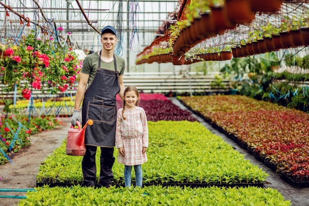 Longitud total de joven trabajador feliz y niña pequeña de pie en el vivero de plantas y mirando a la cámara