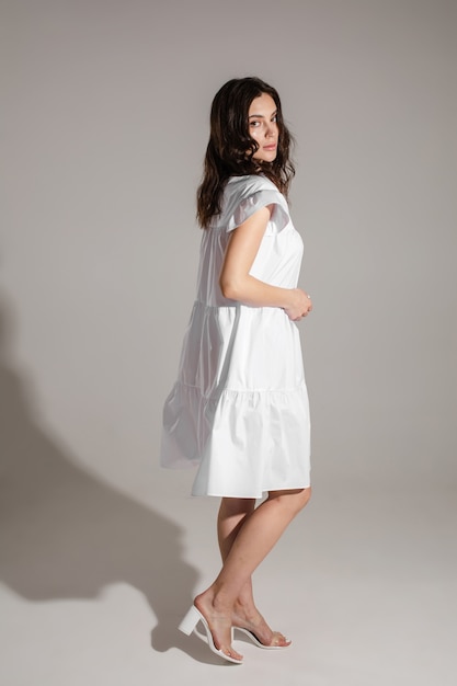Longitud total de una joven tranquila con un vestido blanco y mirando a la cámara mientras posa en el estudio. Moda. Compras. Concepto de verano