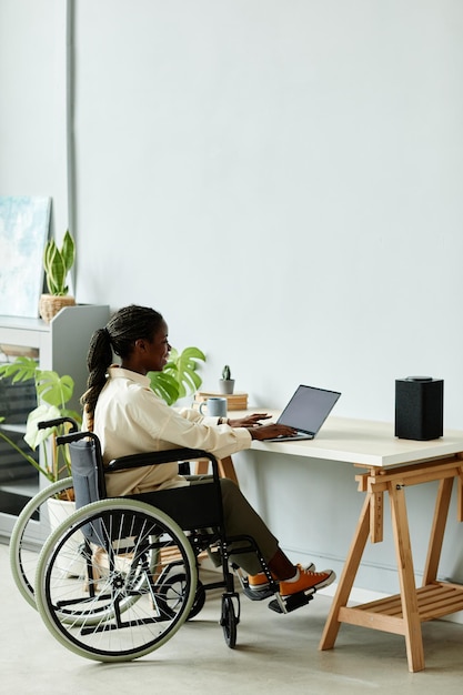 Longitud total mínima de una joven negra en silla de ruedas que usa una computadora portátil mientras trabaja en el espacio de copia de la oficina en casa