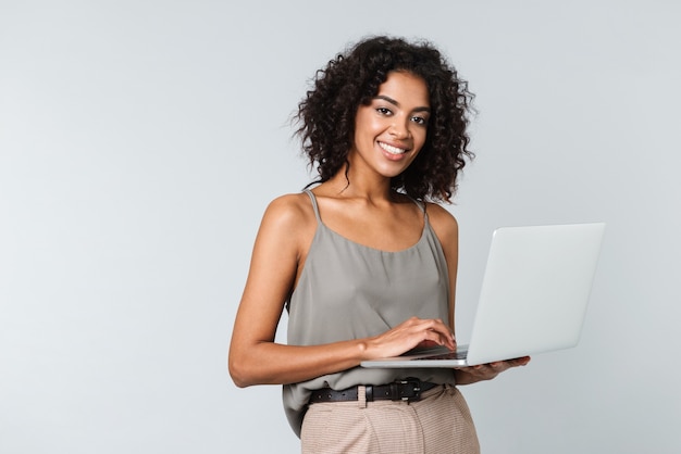 Longitud total de una mujer africana joven feliz vestida casualmente que se encuentran aisladas, trabajando en equipo portátil