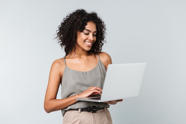 Longitud total de una mujer africana joven feliz vestida casualmente que se encuentran aisladas, trabajando en equipo portátil