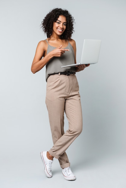 Longitud total de una mujer africana joven feliz vestida casualmente que se encuentran aisladas, trabajando en equipo portátil