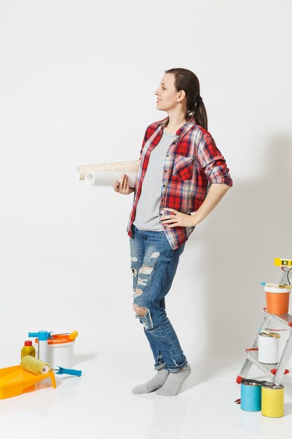 Longitud total de mujer bonita en ropa casual de pie cerca de instrumentos para el apartamento de renovación aislado sobre fondo blanco. Papel pintado, accesorios para pegar, herramientas de pintura. Concepto de reparación.