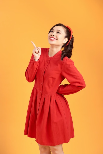 Longitud total de mujer coqueta con sonrisa radiante vistiendo vestido rojo con la mano en la cadera aislado sobre fondo amarillo