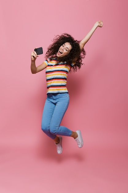 Longitud total de mujer divertida de 20 años con cabello rizado sosteniendo teléfono inteligente y tomando foto selfie aislado en rosa