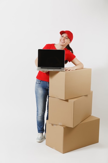 Longitud total de mujer de entrega en gorra roja, camiseta aislada sobre fondo blanco. Mensajero femenino cerca de cajas de cartón vacías, computadora portátil, pantalla vacía negra en blanco. Recibiendo paquete. Copie el espacio.