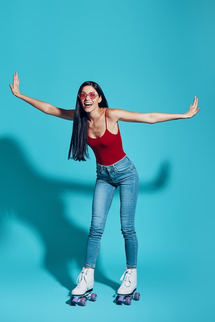 Foto longitud total de mujer joven atractiva y sonriendo mientras patina contra la pared azul