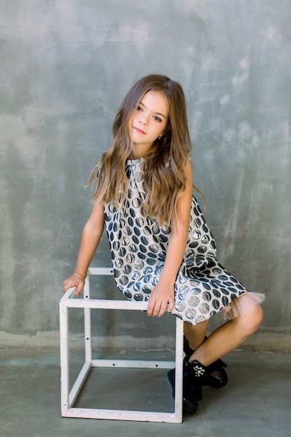 Longitud total de niña encantadora encantadora en vestido gris de moda sentado en el cubo blanco