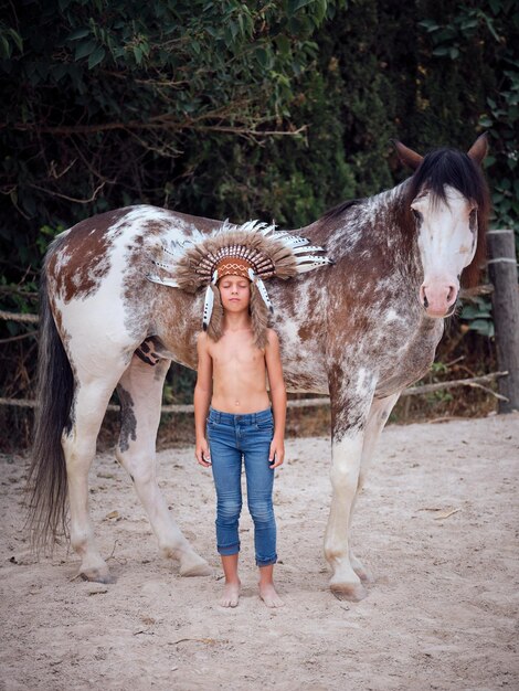 Longitud total de un niño juguetón vestido con cucarachas y jeans de pie con los ojos cerrados cerca de un caballo blanco con manchas en el paddock del rancho