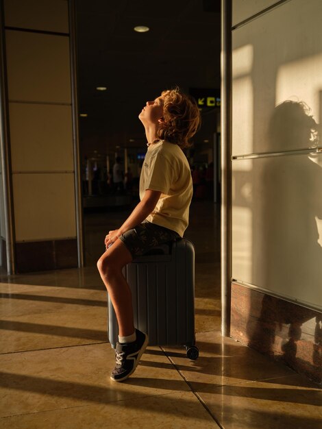 Longitud total de un niño sentado en una maleta y esperando la salida en el aeropuerto mientras viaja en vacaciones de verano