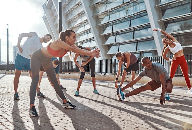 Longitud total de personas en ropa deportiva calentando y estirando mientras hacen ejercicio en la acera al aire libre