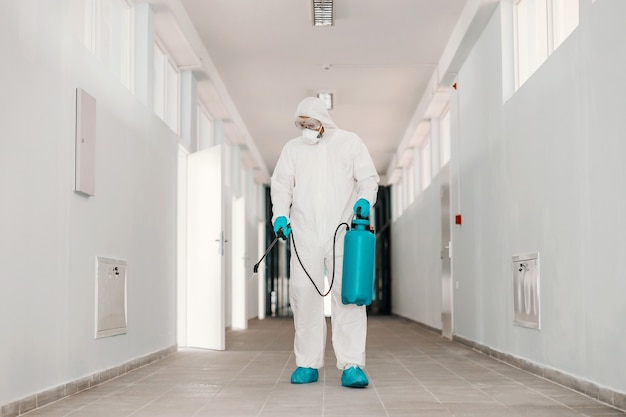 Longitud total de trabajador en uniforme estéril, con mascarilla sosteniendo el rociador con desinfectante y rociando el pasillo de la escuela.