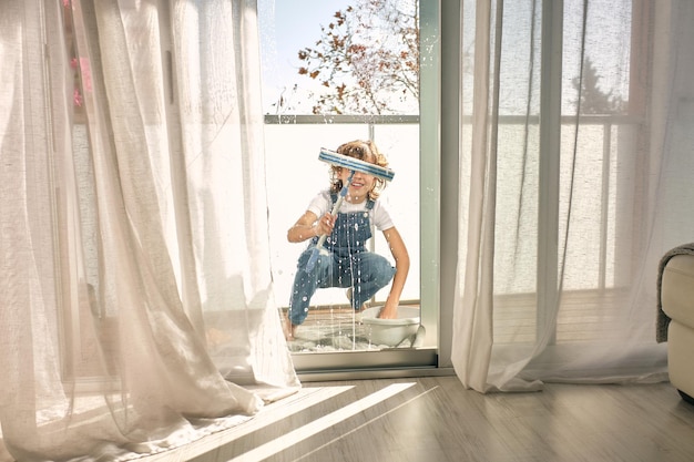 Longitude completa de menino sorridente em roupas casuais agachado enquanto faz tarefas domésticas e lava janelas com esfregão na varanda em casa