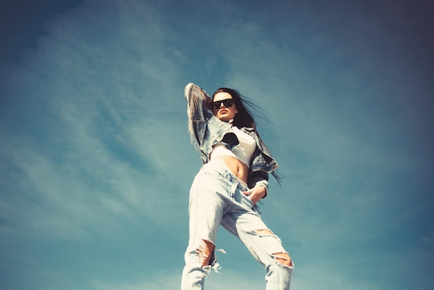 Look de alta moda glamour linda sexy e estilosa caucasiana jovem morena modelo em roupas de jeans hipster posando no fundo do céu azul