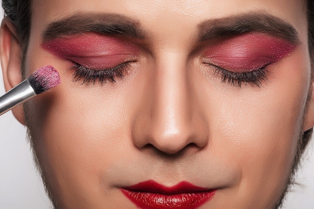 Look de maquillaje para hombres. Retrato de primer plano de un joven barbudo con un pincel de maquillaje