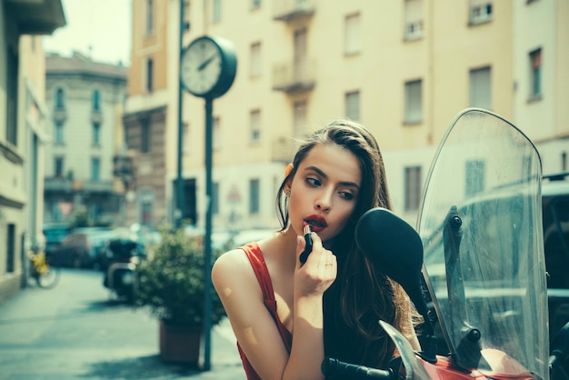 Look de moda de la calle. Aplicar maquillaje al caminar. Chica de moda sexy con labios rojos pone lápiz labial mirando en un espejo de motocicleta.