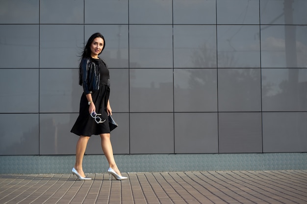 Look de moda callejera. Bastante joven con elegante vestido negro y chaqueta de cuero con flecos negro de moda con elegantes gafas de sol en la mano. Muro urbano en espacio de copia.