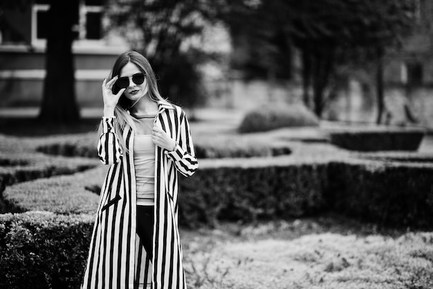 Look de mujer de moda con pantalones de cuero de chaqueta de traje a rayas blancas y negras y gafas de sol posando contra arbustos en la calle Concepto de chica de moda
