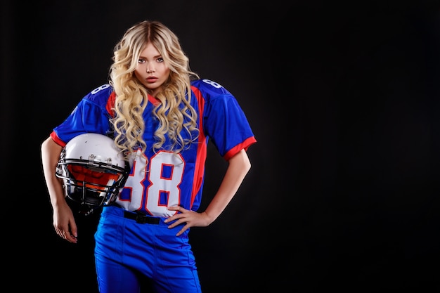 Louro atlético que levanta como a menina do futebol americano no fundo preto. jovem mulher bonita que veste a bola superior da terra arrendada do futebol americano. foto de estúdio de mulher de futebol americano