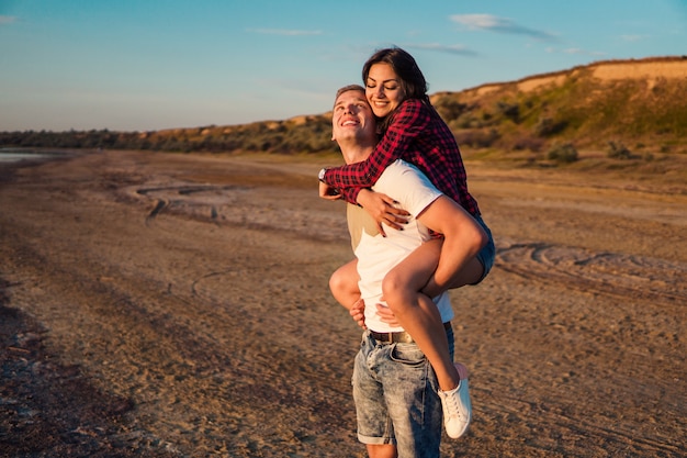 Lovestory de la joven pareja hermosa que lleva a cuestas en la playa al atardecer